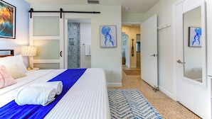Une chambre d’hôtel avec un lit, un ventilateur de plafond, une salle de bain avec un rideau de douche et un couloir menant à d’autres chambres.