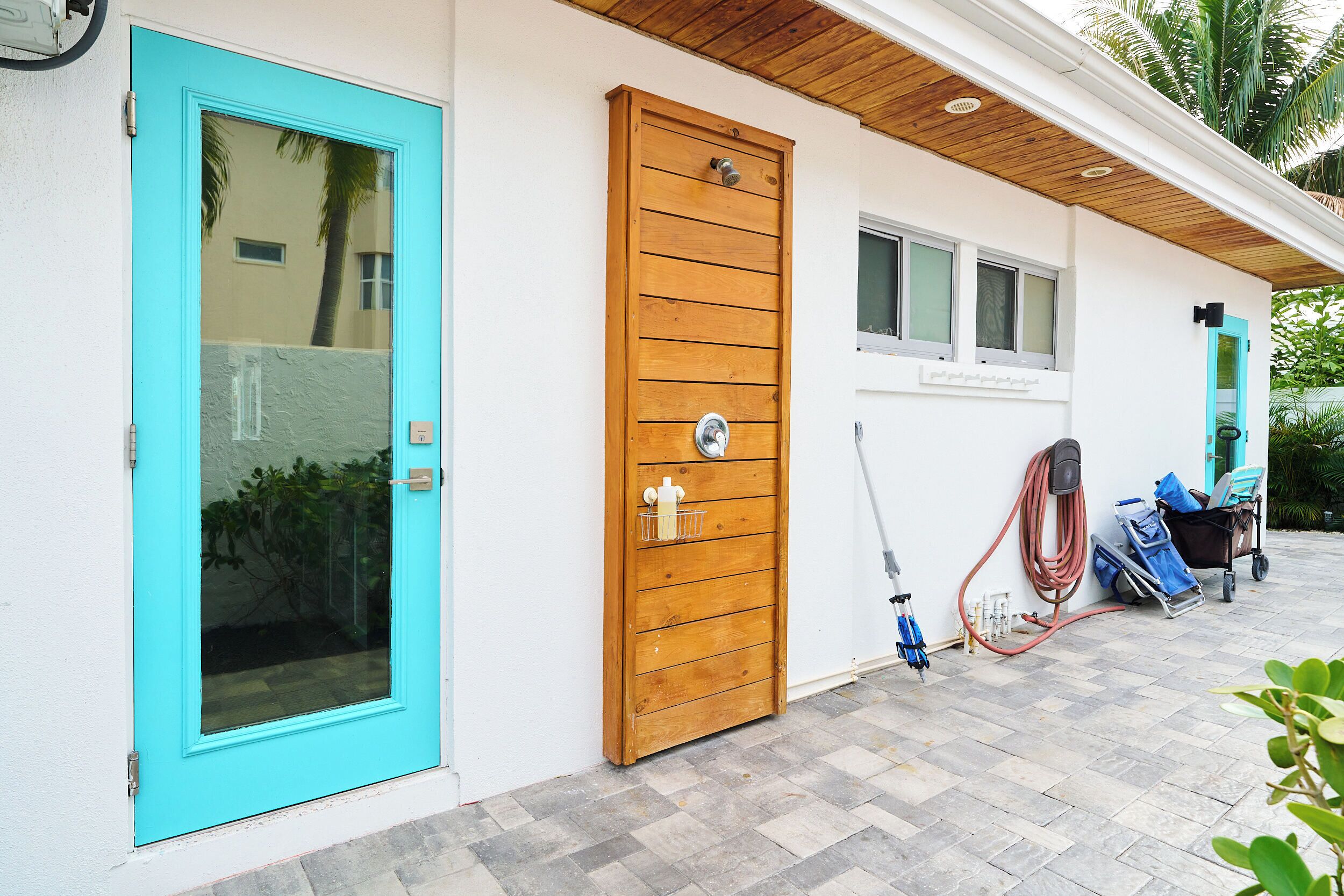 Een gebouw met een turquoise deur, een houten deur en een raam. Een tuinslang en een kar met spullen staan op de grond.