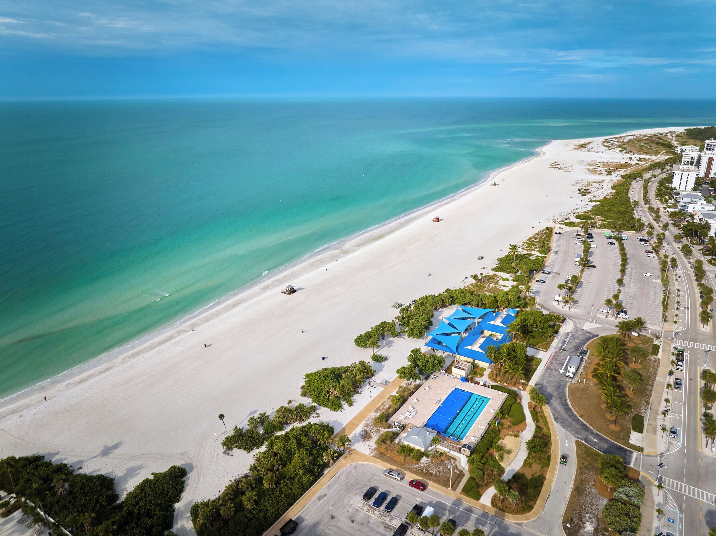Vue aérienne d’une propriété située en bord de mer, dotée d’une piscine, d’un espace de stationnement et d’une route adjacente.