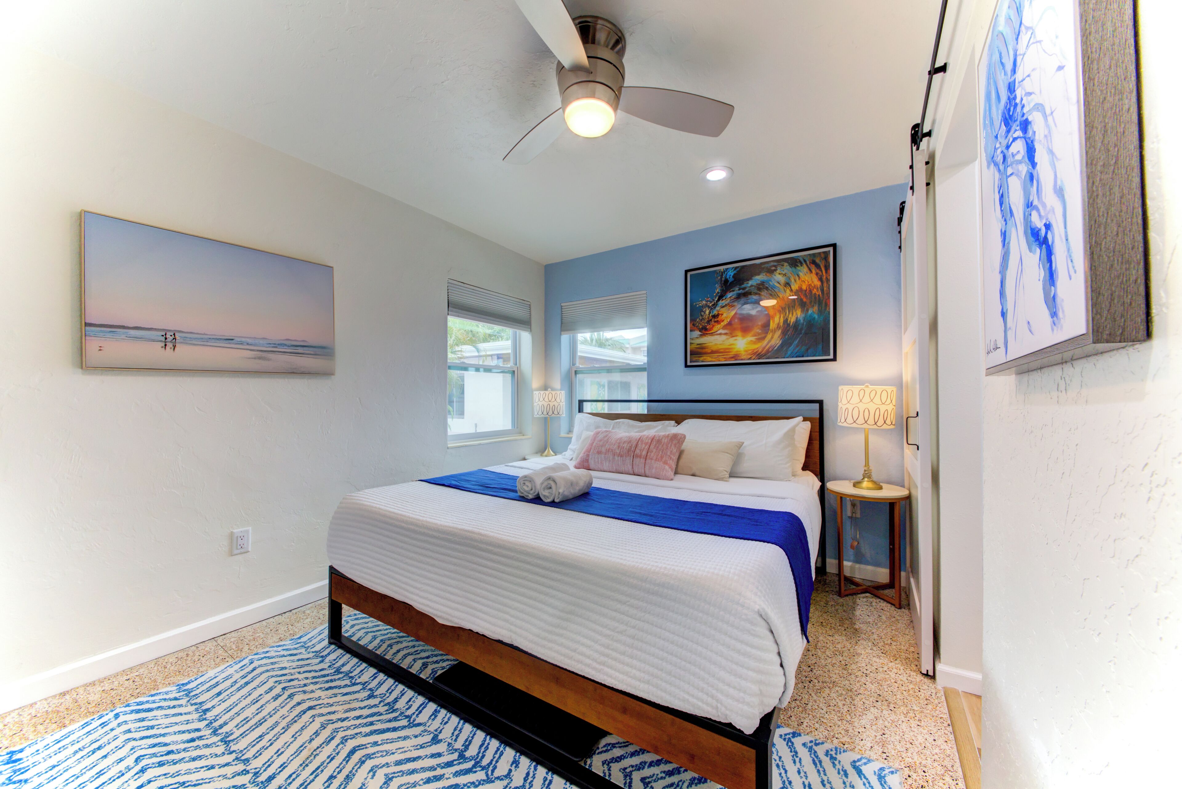A bedroom with a bed, a ceiling fan, a window, and a painting on the wall.