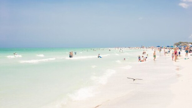 Une plage à l’eau turquoise et limpide, bordée de sable blanc, et où il y a beaucoup de monde.