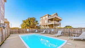 A rectangular pool with a wooden deck, surrounded by a wooden fence. There are lounge chairs and a wooden deck with railings.