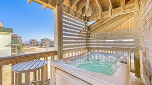 A wooden deck with a hot tub and a small table, overlooking a beach with houses.
