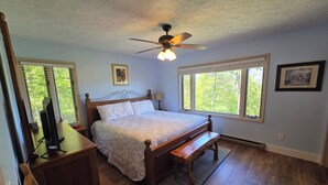Quarto com cama de madeira, um ventilador de teto, escrivaninha com cadeira e duas janelas com persianas.