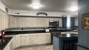 A kitchen with light wood cabinets, a black countertop, a stainless steel microwave, a stove, and a bar area.