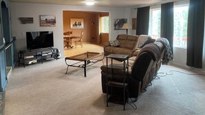 A living room with a sofa, TV, and a glass-top coffee table. There is a dining area in the background with a table and chairs.