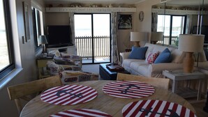 A cozy living room with a large TV, a white sofa with blue cushions, and a wooden table with nautical-themed placemats. - Gorgeous 2 bed 2 bath beachfront condo (North Myrtle Beach)