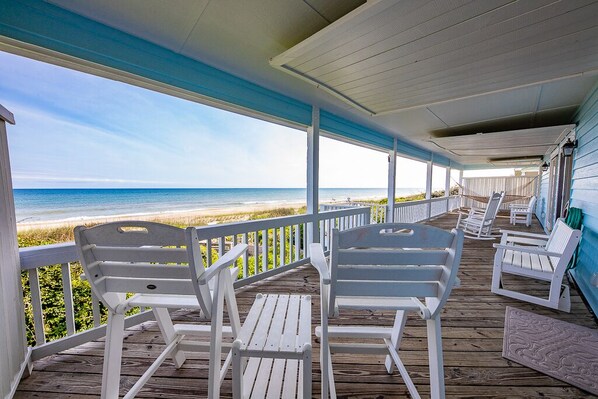 A covered deck with white chairs and a view of the beach.