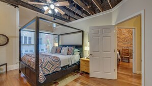 A four-poster canopy bed with a patterned bedspread, a wooden nightstand with a lamp, and a view of a brick wall through an open door.