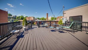 A wooden deck with outdoor seating, including tables and chairs, overlooking a cityscape with buildings and a clear sky.