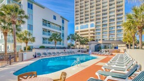 Ein Hotelpoolbereich mit Liegestühlen und einem Tisch, umgeben von Palmen und Gebäuden.