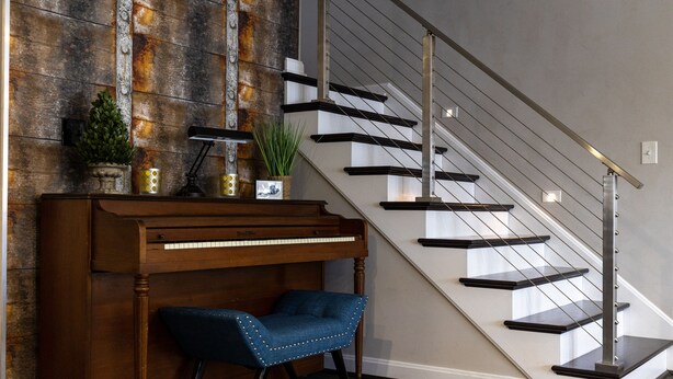 Un piano de cola, un sillón tapizado en azul y una escalera moderna con barandillas de cristal y peldaños de madera.