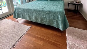A bedroom with a bed, a ceiling fan, a window with blinds, a small table with books, and a door leading to a balcony. - SURFERS DREAM STUDIO (Waianae)