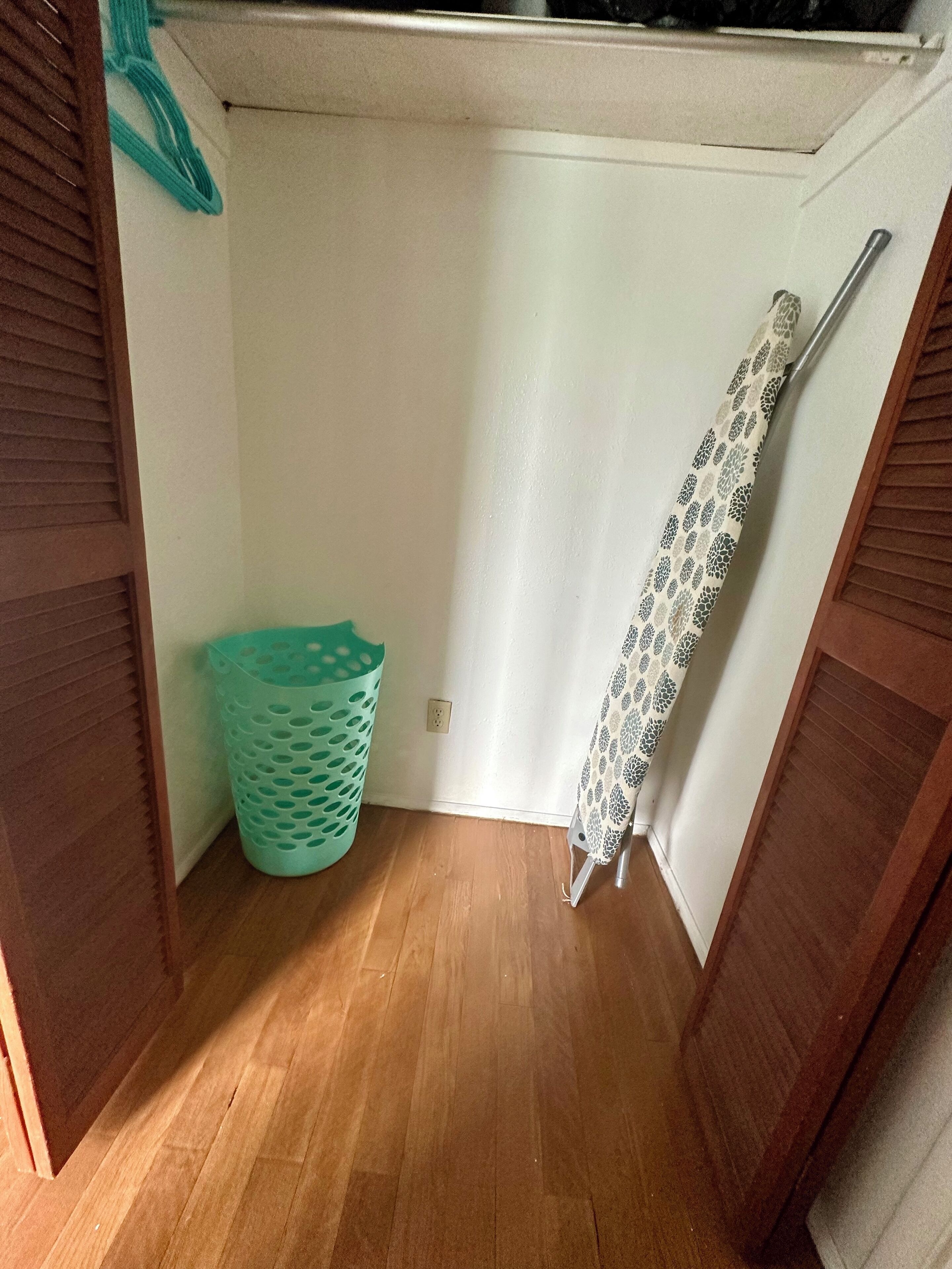 A small laundry room with a wooden floor, a green laundry basket, and a patterned shower curtain.