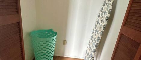 A small laundry room with a wooden floor, a green laundry basket, and a patterned shower curtain.