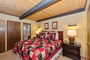 Un dormitorio con una cama grande, vigas de madera en el techo, dos mesitas de noche con lámparas y una puerta a la izquierda.