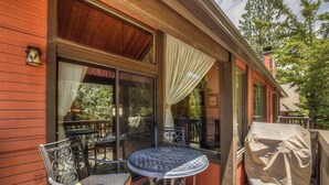A covered outdoor seating area with metal chairs and a round table, a covered grill, and a wooden deck. - Walk to Snow Summit! National Forest & Ski Slope Views! Close to Hiking Trails! (Big Bear Lake)