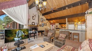 A cozy living room with a fireplace, comfortable seating, a television, and a kitchen area. - Walk to Snow Summit! National Forest & Ski Slope Views! Close to Hiking Trails! (Big Bear Lake)