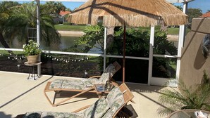 Un espacio de estar al aire libre con techo, amueblado con mobiliario de mimbre y una sombrilla de paja, con vistas a un jardín ajardinado y palmeras.