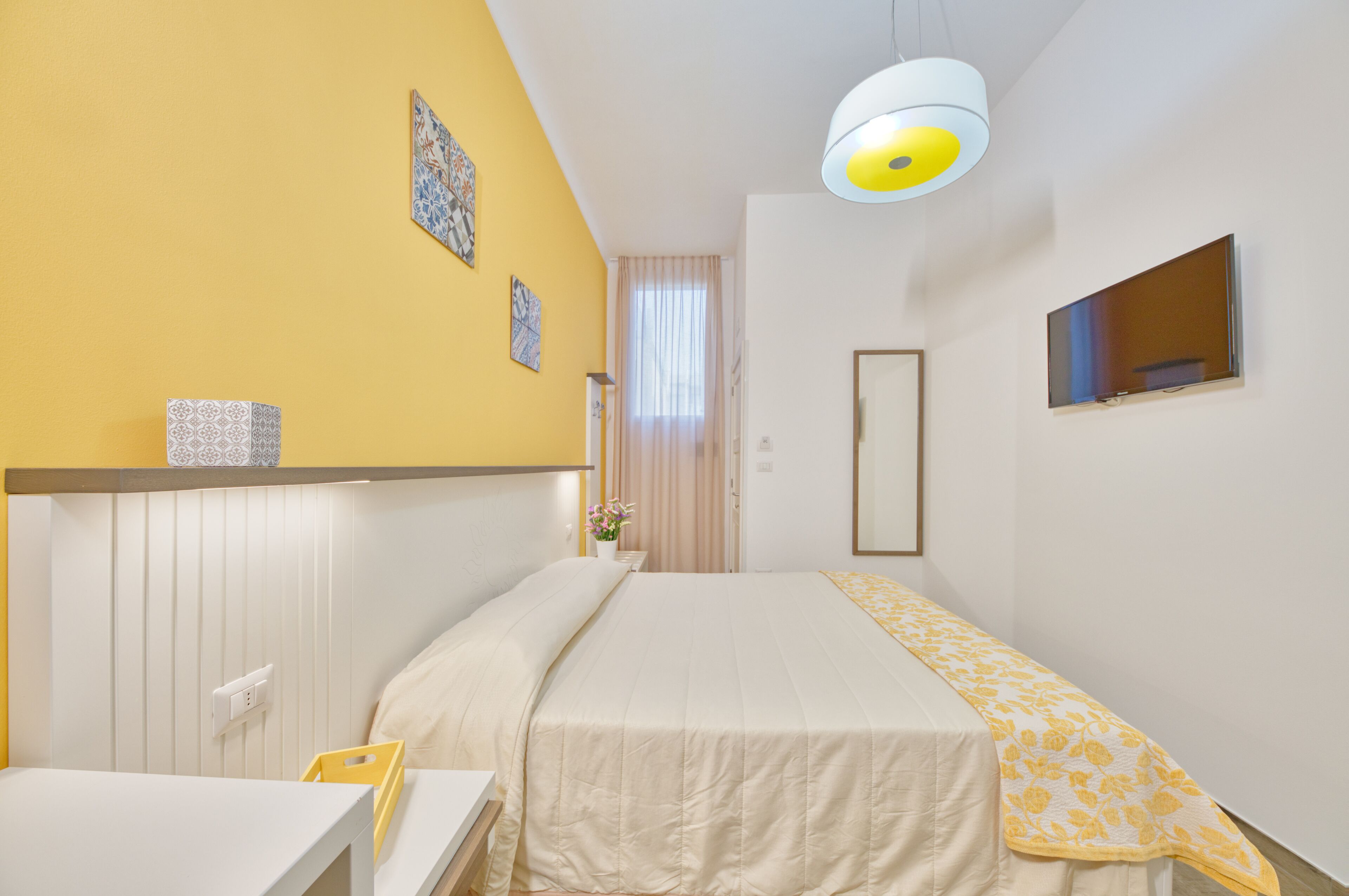 Quarto de hotel com cama, escrivaninha, cadeira, mesa de cabeceira, espelho e janela com cortinas.