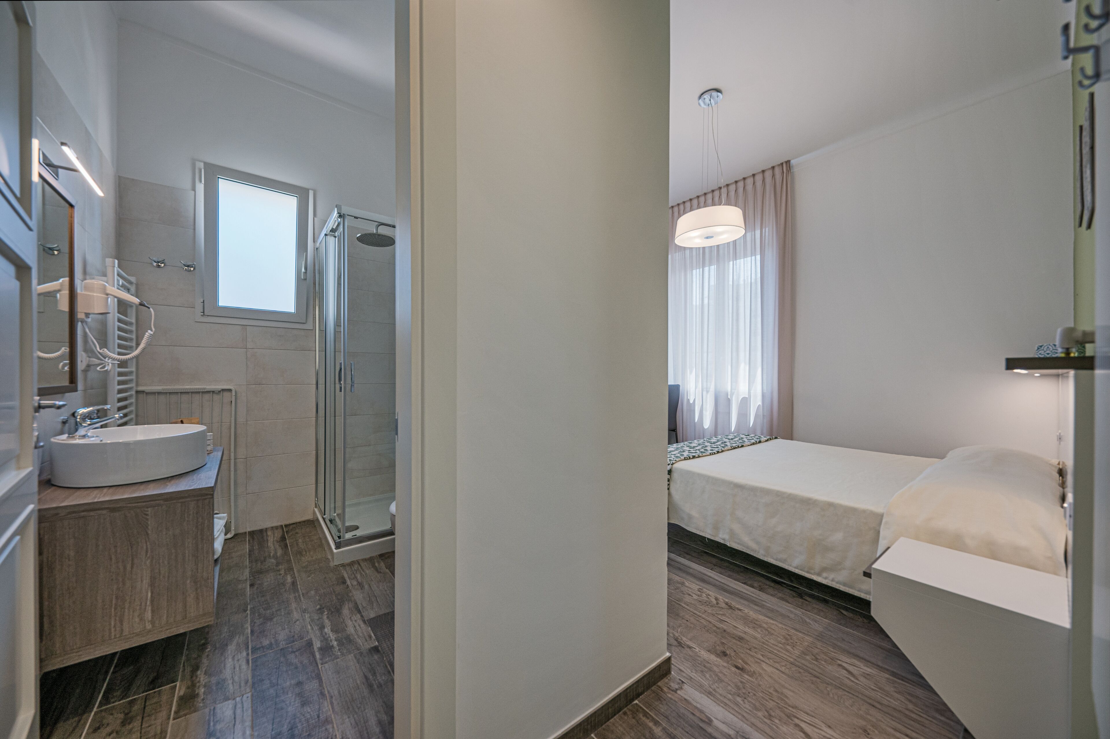 A modern hotel room with a bathroom featuring a sink, mirror, and shower. The room includes a bed with a bedside table and a window with curtains.