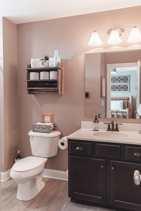 A bathroom with a toilet, a vanity with a mirror, and a shelf above the toilet. - Hocking Hills |Hot Tub, Game Room & Private Trails (Rockbridge)