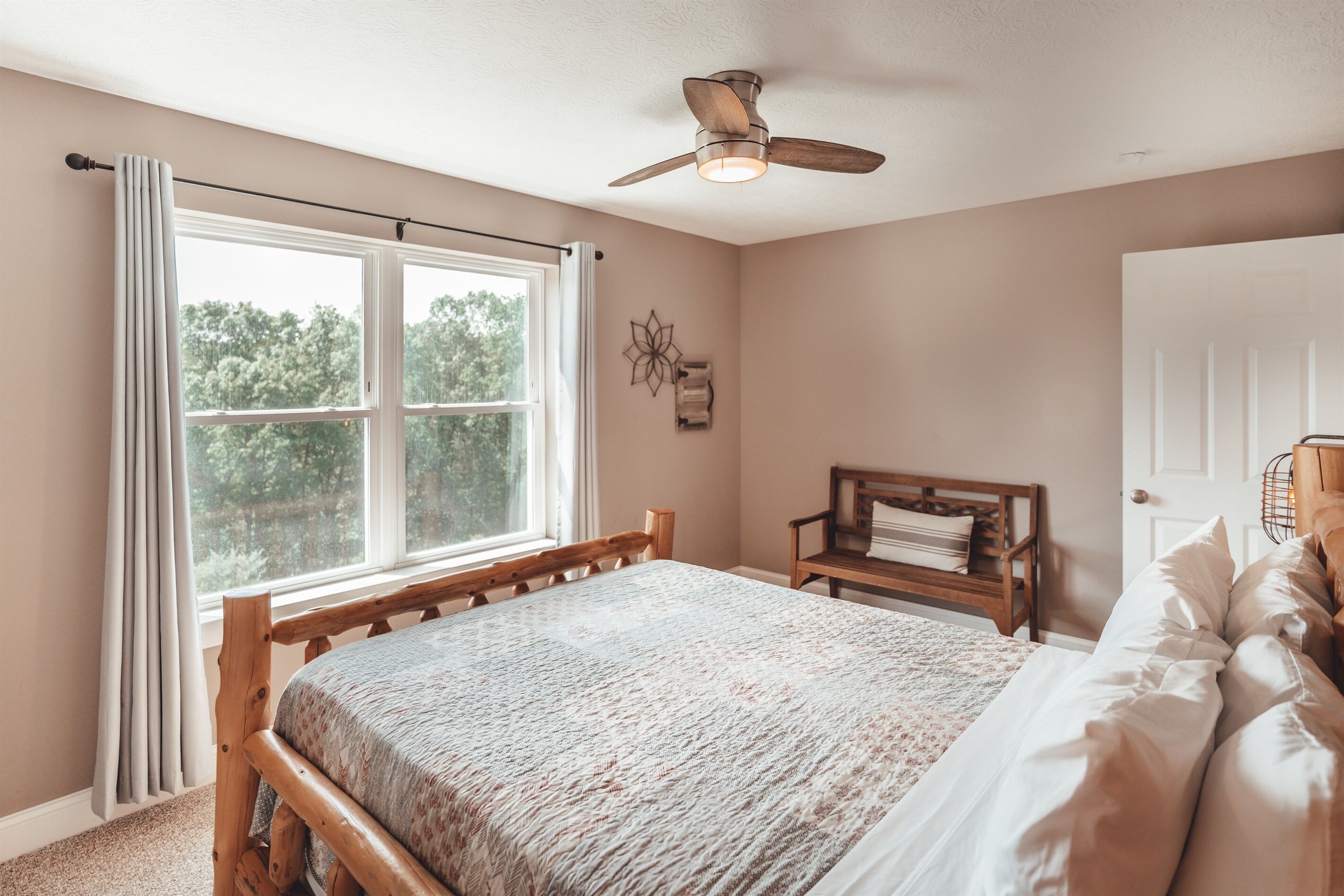 Een slaapkamer met een houten bed, een plafondventilator, een raam met gordijnen, een nachtkastje en een deur.