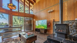 A cozy living room with a stone fireplace, a large TV, wooden walls, and a view of the outdoors. - Cub's cabin Family house inside the gates of Yosemite Park!!! (Yosemite National Park)