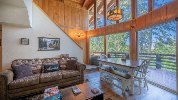 A cozy living room with a wooden ceiling, a sofa with patterned cushions, a coffee table with books, and a dining table with chairs. - Cub's cabin Family house inside the gates of Yosemite Park!!! (Yosemite National Park)