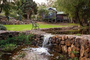 Una cabaña rústica con una cascada de piedra, rodeada de exuberante vegetación y una pequeña terraza de madera.