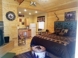 A log cabin room with a bed, a small table, a chair, a clock, and a kitchen area.