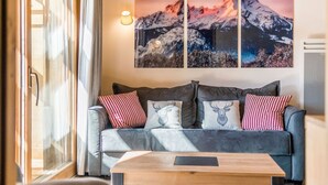 Ett modernt vardagsrum med en grå soffa, röda och vita mönstrade kuddar och ett soffbord i trä. En stor tavla med ett berglandskap, som hänger på väggen, syns tydligt.