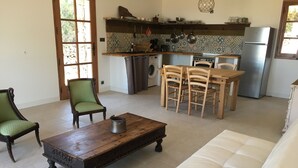 Een woonkamer met een bank, een houten salontafel en twee stoelen. Er is een keukenhoek met een koelkast, fornuis en diverse keukenbenodigdheden.