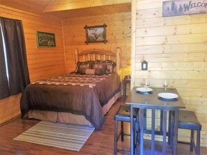 A wooden cabin interior with a bed, a table with two wine glasses, and a welcome sign.
