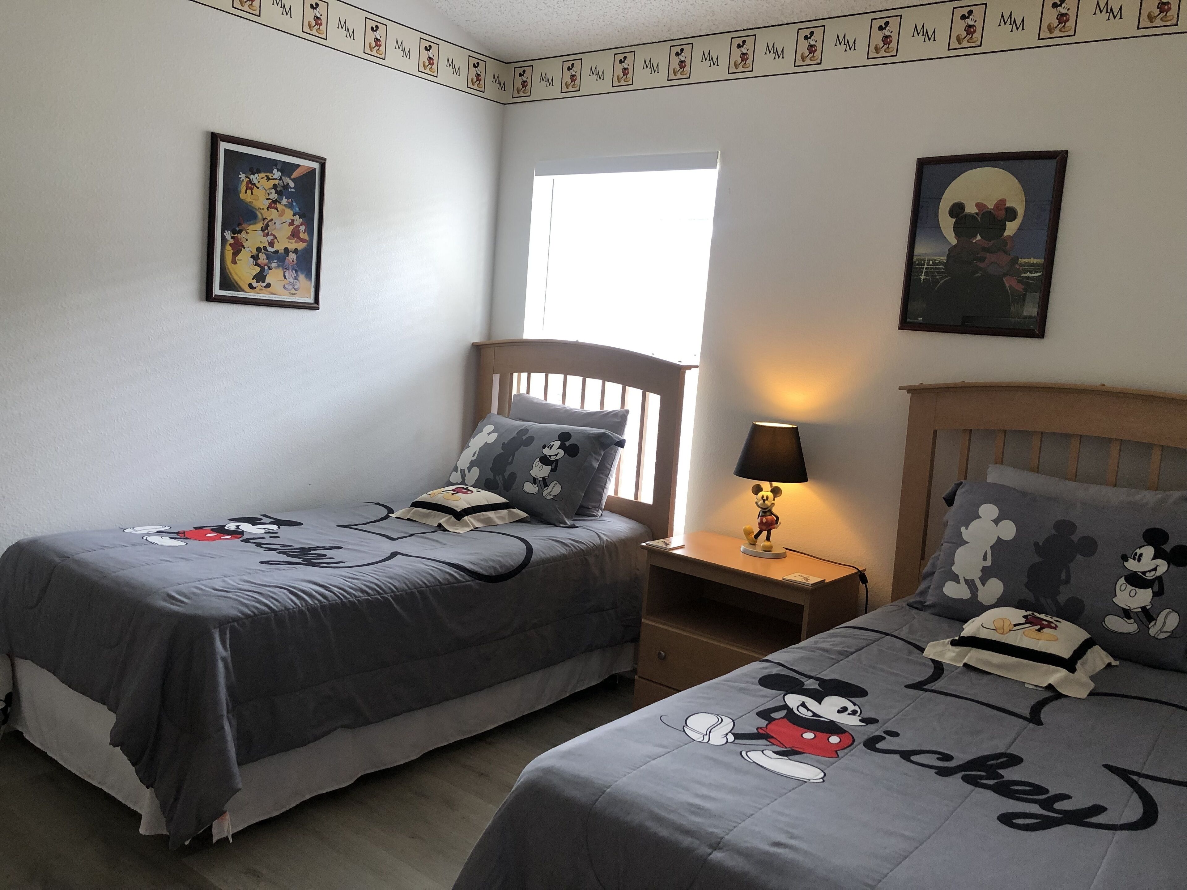 A bedroom with two beds, each with Mickey Mouse-themed bedding. There is a window providing natural light, a nightstand with a Mickey Mouse lamp, and wall decorations featuring Mickey Mouse.