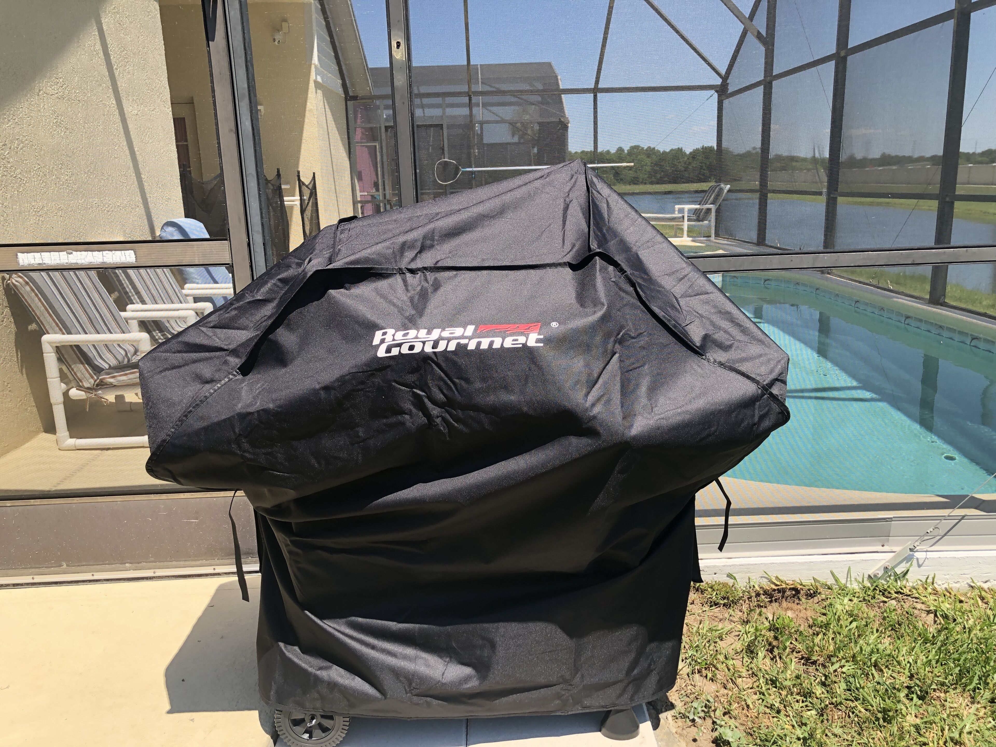 Una parrilla para barbacoa cubierta con una funda negra de la marca Royal Gourmet, ubicada cerca de una piscina con vistas a un lago y sillas al fondo.