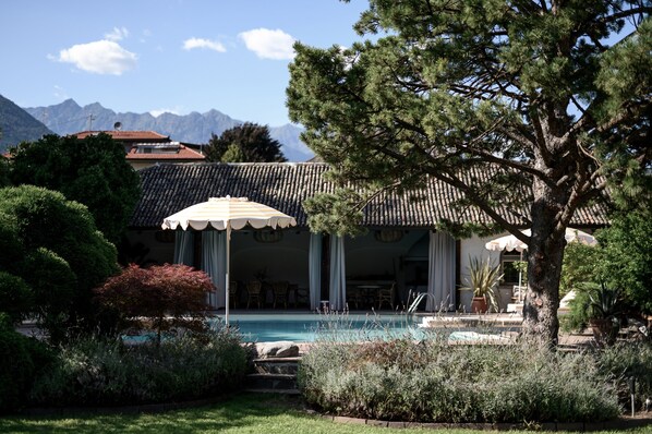 A pool area with a covered seating area, surrounded by lush greenery and a mountain backdrop. - Villa Arnica (Lana)