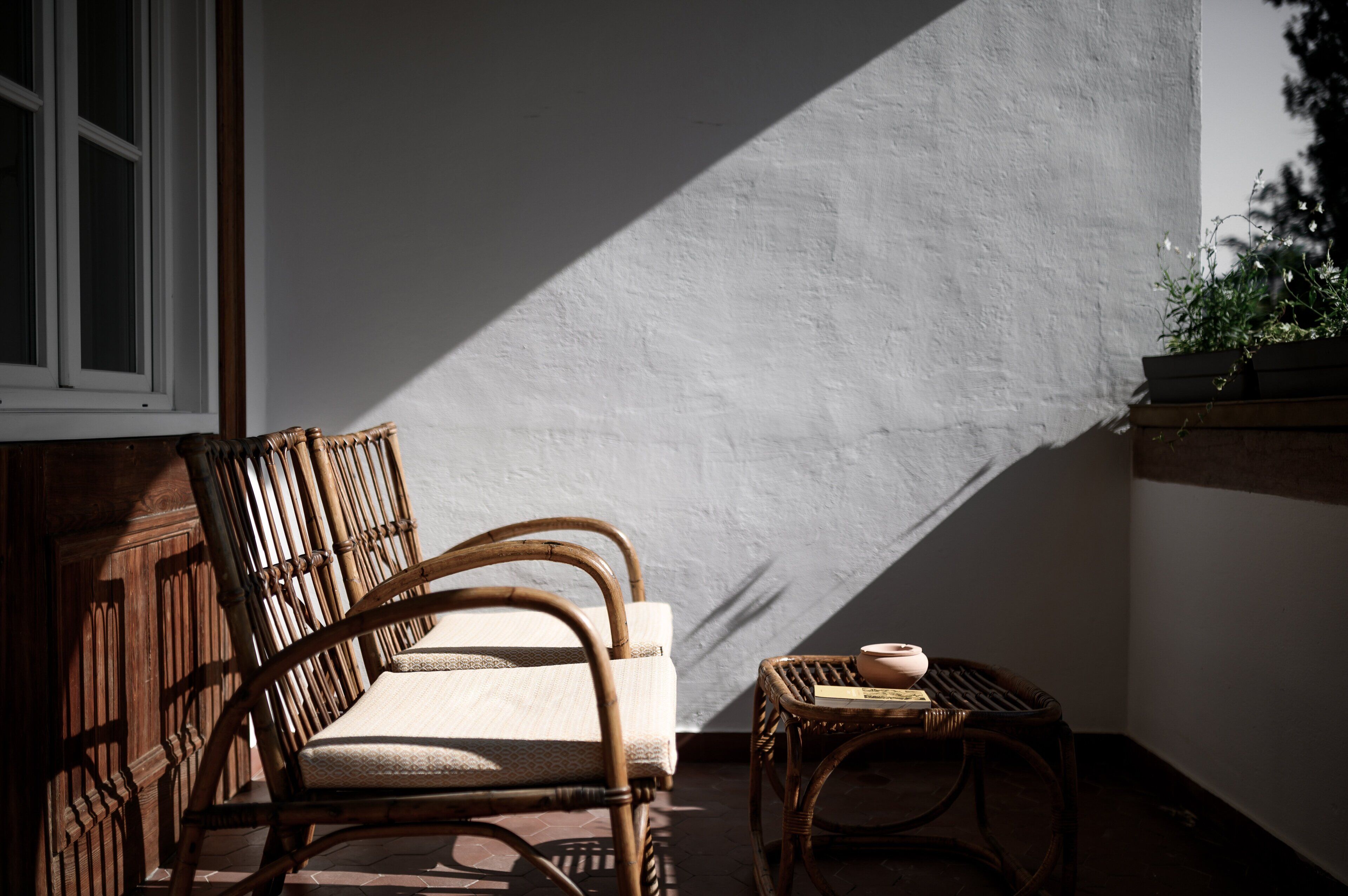 A wicker chair and a small table with a book and a cup, positioned near a window with a white wall and a shadow cast on it.