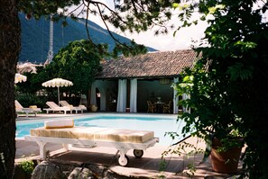 A pool area with lounge chairs and umbrellas, a building with a tiled roof, and a mountainous backdrop. - Villa Arnica (Lana)