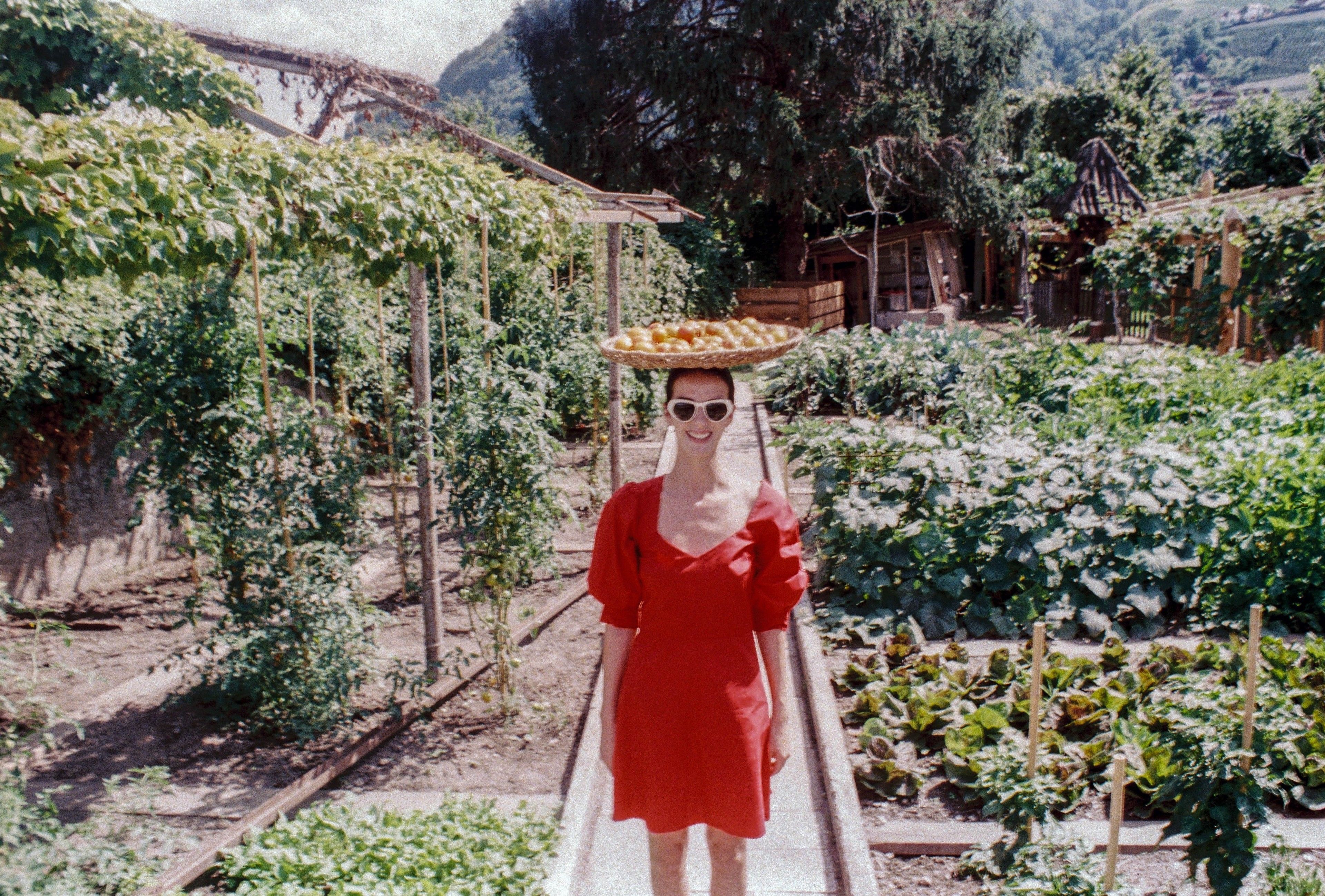 A woman in a red dress walking on a pathway in a garden with various plants and a wooden structure in the background.