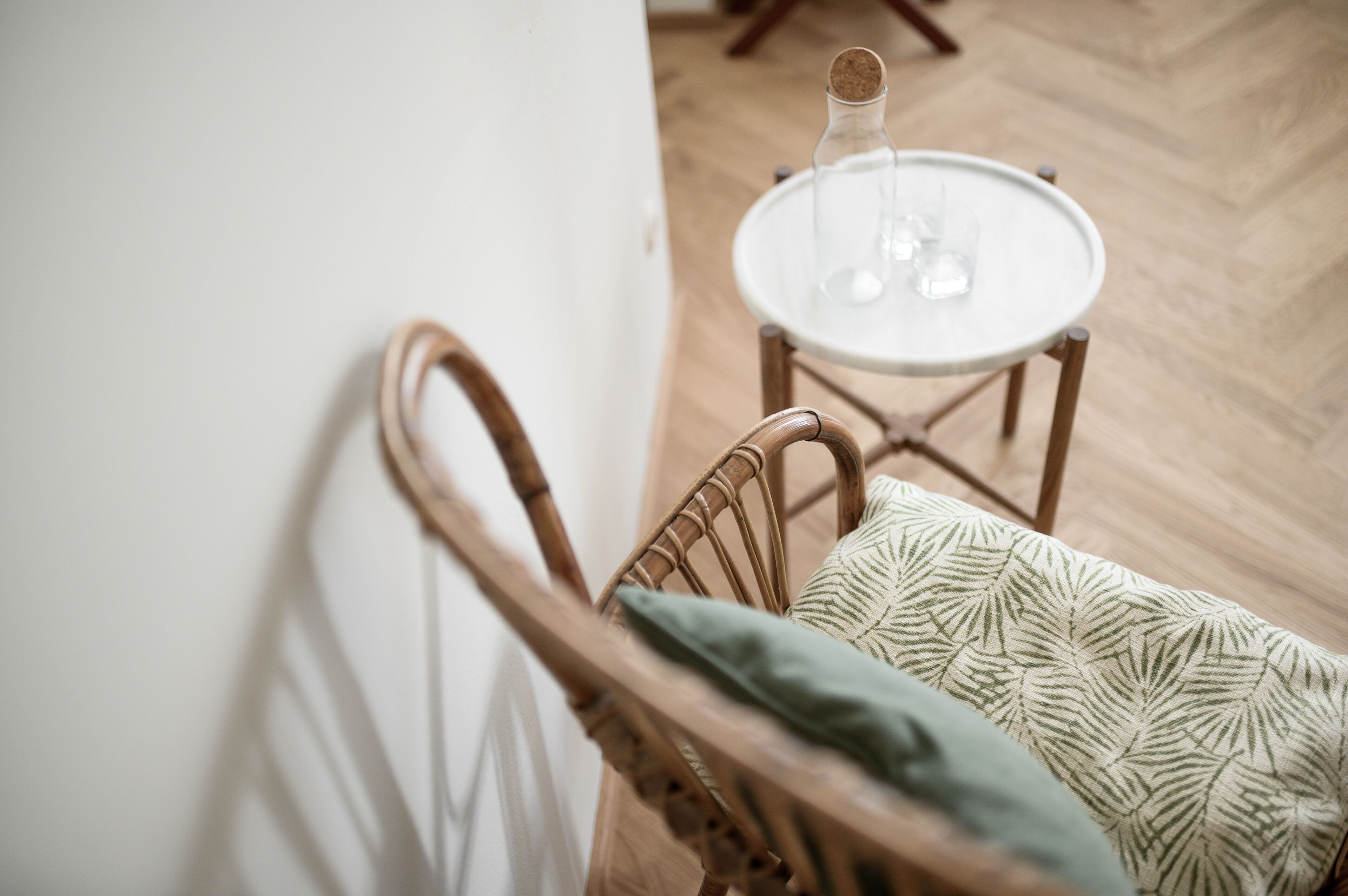 A wicker chair with a patterned cushion and a small round table with a clear glass bottle and glasses.
