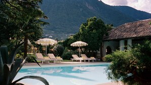 Ein Poolbereich mit Liegestühlen und Sonnenschirmen, umgeben von viel Grün und einem Bergpanorama.