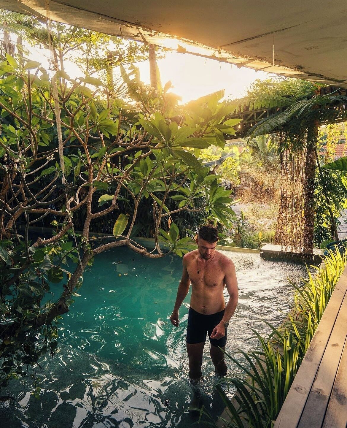 Un homme se tient dans une piscine entourée de plantes tropicales et bordée d’une terrasse en bois.