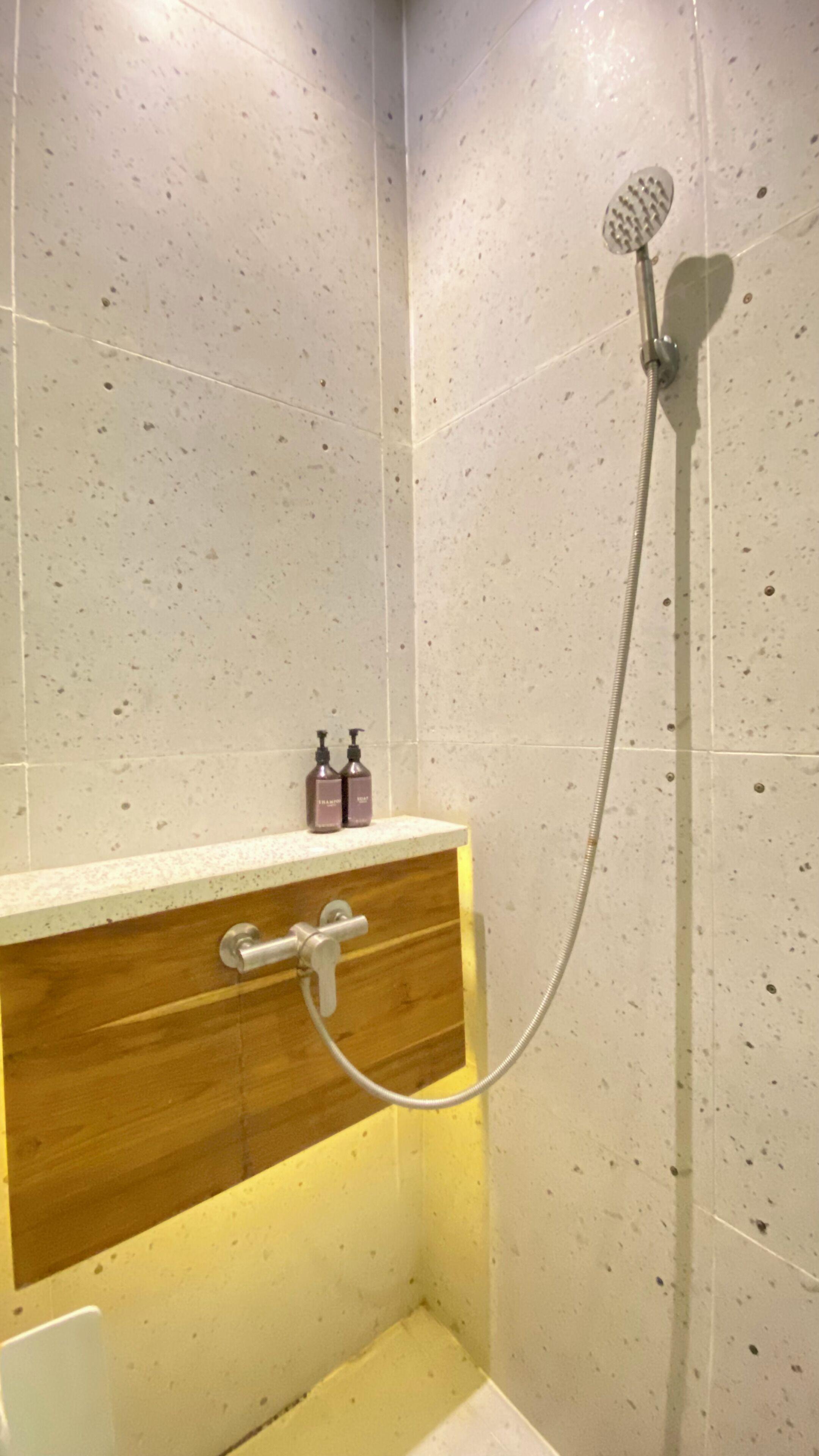 A shower with a handheld showerhead and a wall-mounted faucet. There are two bottles of shampoo on a shelf above the shower.