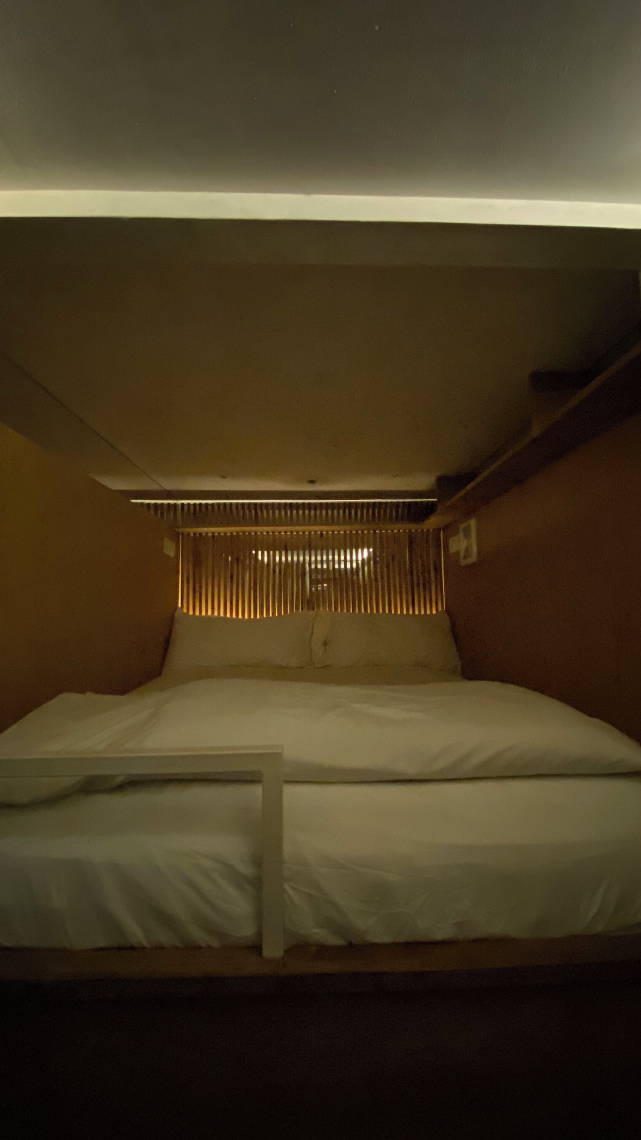 Una cama litera con cabecera de listones, un pequeño estante a la derecha y una ventana con persianas.
