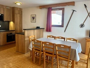 Une cuisine avec des meubles de rangement en bois, une table à manger avec des chaises, un petit réfrigérateur et une fenêtre avec un rideau.