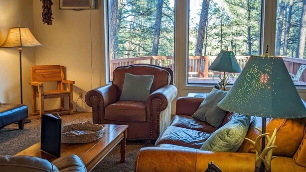 A cozy living room with a leather armchair, a sofa, and a coffee table. There is a lamp on the sofa and a lamp on the coffee table. The room has large windows with a view of trees outside. - Mama Bears Cabin (Ruidoso)