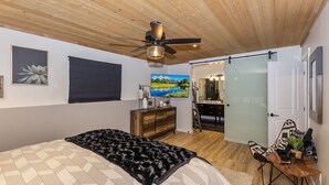 Ein Schlafzimmer mit Bett, Deckenventilator, Kommode, Fernseher und angrenzendem Badezimmer, durch eine Glastür sichtbar.
