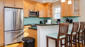 A kitchen with wooden cabinets, stainless steel appliances, a green tiled backsplash, and a bar area with wooden chairs. - Gorgeous Home 1 Block from Ocean and Banks Channel (Wrightsville Beach)