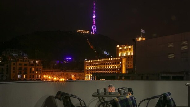 Eine Dachterrasse mit Tisch und Stühlen, mit Blick auf eine Skyline mit beleuchteten Gebäuden und einem markanten Turm.